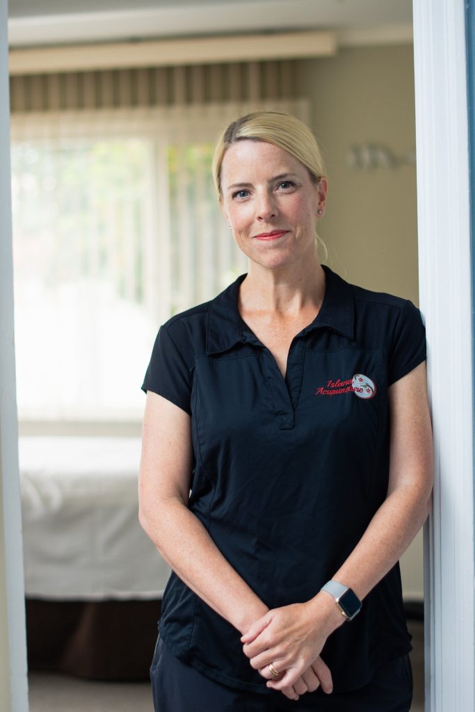 A grinning adult leans against the door of an acupuncture treatment room with open blinds and sunlight coming through. 